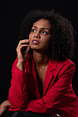 Pensive young Hispanic female model with Afro hairstyle in red coat looking away while sitting against black background