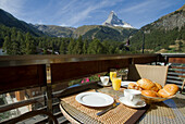 An outdoor dining setup overlooking the majestic Matterhorn mountain, with a spread of breakfast items including orange juice, croissants, and coffee surrounded by lush greenery
