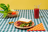 Appetizing healthy sandwich with fresh salad served on plate with olives near glass of tomato juice with glass straw on checkered tablecloth