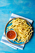 Top view of ceramic plate with French fries and bowl of sour sweet sauce placed on striped napkin against blue background