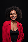 Smiling young Hispanic female model with Afro hairstyle in red coat looking at camera while standing against black background