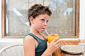Side view of thoughtful cute boy drinking mango juice from glass and looking away seriously while sitting at table in cafe during summer vacation