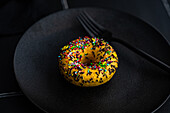 High angle of sweet banana donut with colorful sprinkles placed on black plate with fork against dark surface