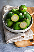Raw feijoa fruits, also known as pineapple guavas, displayed in a ceramic bowl on a wooden cutting board with a linen cloth.