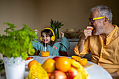 Senior man eating orange and happy cute girl with headphones listening music and dancing while drinking juice with straw at table
