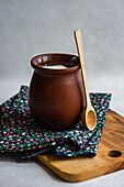 Traditionelle Portion georgischer saurer Joghurt, bekannt als Matsoni, in einem Tontopf neben einem Holzlöffel auf einer bunten Serviette auf einem Holztablett vor einem unscharfen Hintergrund