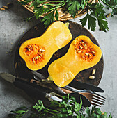 Kürbishälften auf rundem Holzschneidebrett mit Besteck auf grauem Küchentisch mit Kräutern. Kochen Vorbereitung zu Hause mit saisonalen Herbst Kürbis. Gesundes Kochen. Ansicht von oben