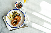 Top view of well-prepared grilled trout steak with capers and lemon served on a white plate with a blue rim placed on striped napkin, cutlery, glass of water and bowl with capers