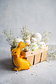 A plush yellow Easter bunny gazes at a basket filled with decorated white eggs and sprigs of baby's breath on a textured grey background