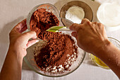 Top view of Hands of crop anonymous person pouring cocoa powder into a bowl with other baking ingredients for a chocolate dessert on table