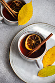 Top view of warm cup of spiced tea garnished with cinnamon sticks, anise and dried orange slices complemented by vibrant yellow autumnal leaves on a light gray surface