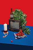 High angle of vintage television set surrounded by an array of objects, including a bottle with a striped label, fresh grapes on a checkered cloth, a white candle and green tinsel, all set against a red backdrop on a blue table