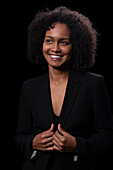 Positive young Hispanic female in elegant outfit smiling and looking away while standing against black background