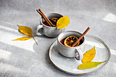 A warm cup of spiced tea garnished with cinnamon sticks, anise and dried orange slices complemented by vibrant yellow autumnal leaves on a light gray surface