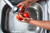 High angle of anonymous African American male chef washing fresh ripe red tomatoes in chrome sink with water tap while preparing ingredients for cooking process in kitchen