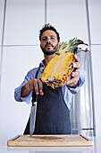 Low angle of Confident adult male chef in apron looking at camera while standing at chopping board with knife in hand and showing sliced pineapple against wall with reflecting fridge door in daylight