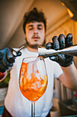 Concentrated young professional barman pouring orange drink from jigger into a glass while preparing refreshing cocktail and standing at counter in bar