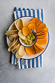 Top view of plate of assorted crisps including potato, paprika, and cheese flavors, served with a bowl of fresh avocado guacamole on a striped napkin