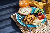 High angle of five ingredients lunch set consists of homemade goat cheese, fresh cucumber and tomato with leaves of basil herb, in addition to fried rice with meat with slices of healthy sour dough bread served with glass of carrot-apple juice against gray background