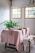 Wooden table with pink tablecloth and dishware placed near bench covered with cloth against potted plant and white brick wall