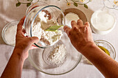 Top view of Hands of crop anonymous person pouring flour into a glass bowl amidst various baking ingredients on a table with a subtle floral tablecloth