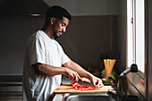 Side view of concentrated young ethnic African American African American male in white t shirt slicing ripe red bell pepper with sharp knife on wooden chopping board while preparing healthy salad in kitchen