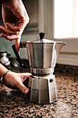 Anonymous young individual hands expertly assembling a stainless steel stovetop espresso maker on a speckled countertop with ambient light filtering through a nearby window