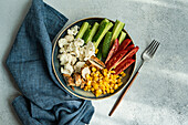 Top view of five ingredients bowl with raw organic vegetables cauliflower, tomato, cucumber, boiled sweet corn and barbeque chicken meat placed on gray surface near napkin and fork