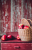 Weidenkorb gefüllt mit frischen, reifen, roten Äpfeln auf einem schäbigen Holzschrank an einer Holzwand in einem Haus auf dem Land während der Erntezeit