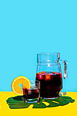 Kaltes Getränk mit Orangenscheibe im Glas und Krug auf Blattdekor über gelbem Hintergrund vor blauem Hintergrund im Studio