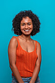 Portrait of friendly African American female fashion model in casual clothes with curly hair smiling while standing against blue background