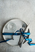 Top view of minimalist yet stylish Easter table setting, with a concrete grey plate bound by a rich blue ribbon, grey cutlery is neatly tied together, while a textured clear glass sits at the center