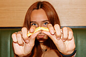 Young woman with red hair and piercing sitting at table and looking at camera while showing french fries in restaurant