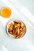 Top view of fried potato with paprika spice served on ceramic plate near orange juice against gray background