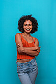 Friendly African American female fashion model in casual clothes with curly hair smiling with crossed arms while standing against blue background