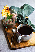 Hoher Blickwinkel auf ein Glas hausgemachtes Kaffee-Eis mit Waffel und frischer Minze neben einem Löffel und einer Tasse Kaffee auf einem Holztablett vor grauem Hintergrund