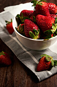 Appetizing strawberries with green leaves placed in white bowl on cloth napkin against wooden table