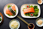 Top view of appetizing fresh salmon fillet with lettuce salad and sushi rolls placed on ceramic plates near chopsticks and seaweed served with soy sauce on table