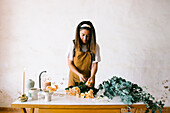 Concentrated female floral designer in apron standing at table and creating floral composition with blooming roses in workshop