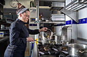Side view of woman in dark uniform putting cut artichokes from bowl on hot frying pan while preparing dish in restaurant kitchen