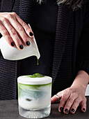 Anonymous female pouring aromatic matcha beverage into glass cup with milk and ice cube while preparing healthy drink