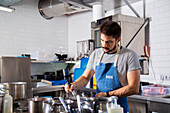 Concentrated young bearded Hispanic male chef in casual clothes and apron stirring boiling dish with whisker in saucepan while cooking in restaurant kitchen in daytime