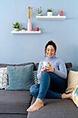 Full body of optimistic barefoot female looking at cup of hot drink while resting on comfortable couch in decorated living room
