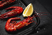 Delicious cooked red prawns on tray with coarse salt and juicy lemon pieces on dark background