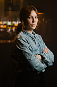Attractive waitress in apron looking at camera with crossed arms while standing near bar counter with bottles of alcohol drinks