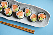 Leckere norwegische Futomaki-Sushi-Rollen mit Lachs und Avocado, serviert mit Stäbchen auf blauem Hintergrund in einem hellen Studio