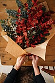 From above of crop unrecognizable female florist standing at wooden table arranging Christmas bouquet in room