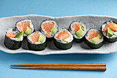 Leckere norwegische Futomaki-Sushi-Rollen mit Lachs und Avocado, serviert mit Stäbchen auf blauem Hintergrund in einem hellen Studio