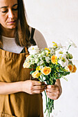 Crop adult Hispanic female florist with dark hair in apron arranging bouquet of assorted gentle fresh flowers against white background