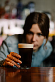 Soft focus of concentrated female bartender looking at glass full of dry stout served on wooden counter in modern bar